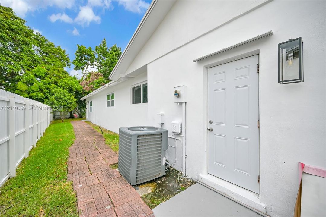 Exterior side profile featuring a white stucco finish, a paved brick walkway, a white vinyl privacy fence, and a modern black-framed wall-mounted lantern