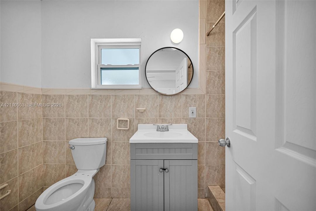 Bathroom featuring neutral-toned wall tile, a gray vanity cabinet with a white countertop, a circular wall-mounted mirror, and a single-hung window