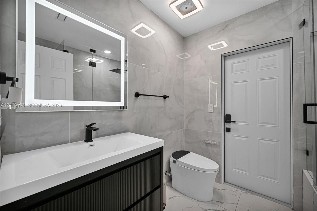 Bathroom featuring a modern vanity with a rectangular integrated sink, wall-mounted matte black faucet, illuminated LED mirror, floor-to-ceiling large-format tile, and a glass-enclosed shower