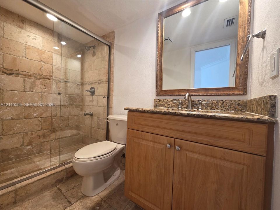 Bathroom featuring a walk-in shower with stone tile surround, glass enclosure, stone-surface countertop, wood-finish cabinetry, and a framed mirror
