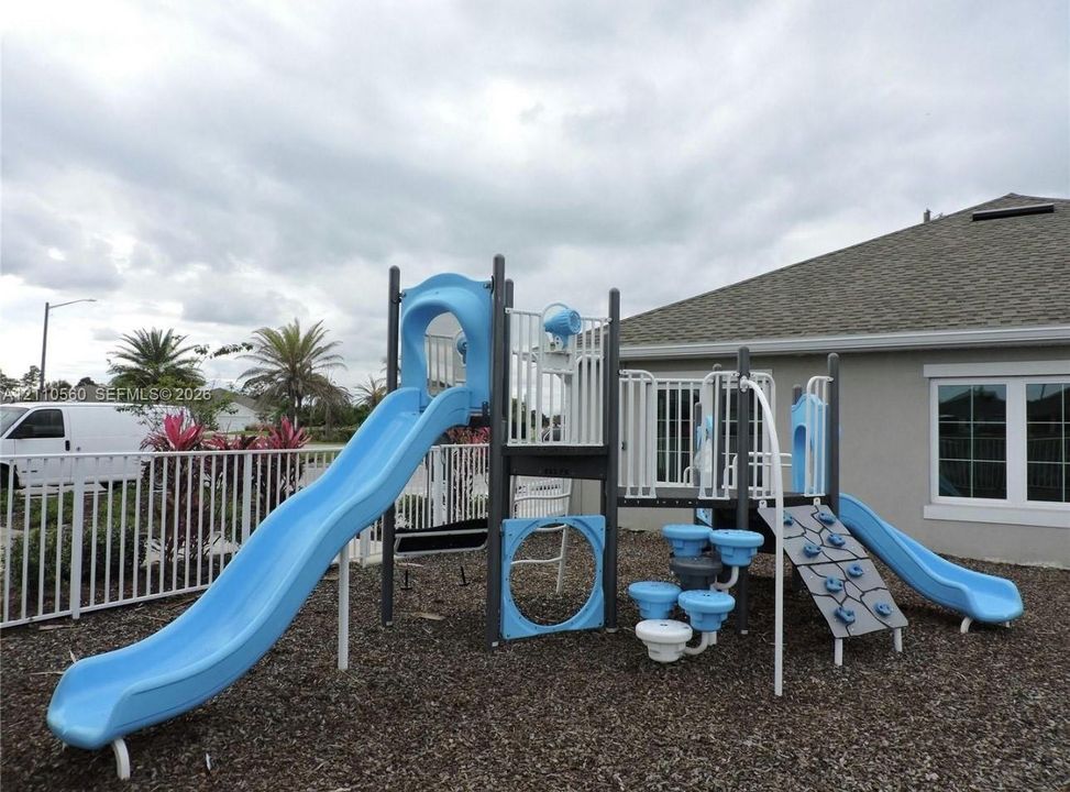 Outdoor playground structure featuring two slides, a climbing wall, and elevated platforms, situated on a ground cover of wood mulch and enclosed by a white vinyl fence