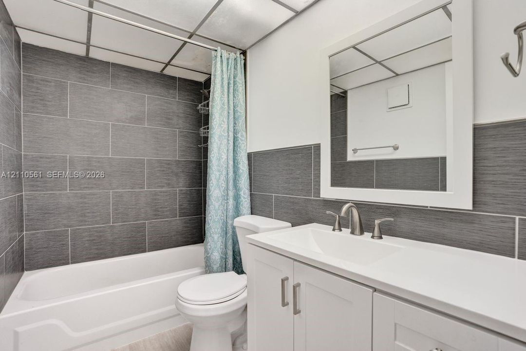 Bathroom featuring a white vanity cabinet with a solid surface countertop, an undermount sink, brushed nickel hardware, and grey textured tile surround in the shower area