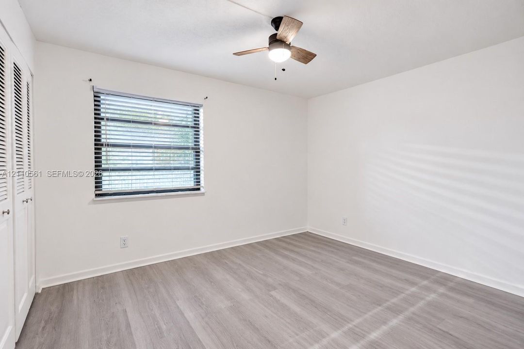 Neutral-toned wood-finish flooring, ceiling fan with integrated lighting, window with horizontal blinds, and double-door closet with louvered panels