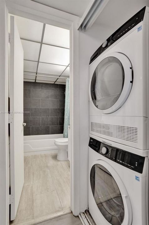 Stacked laundry unit featuring a front-loading washer and dryer, situated near a bathroom with wood-finish flooring, a white bathtub, and dark horizontal wall tiling