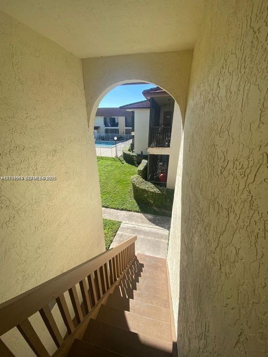 Exterior staircase featuring stucco walls, an arched entryway, and wooden handrail, with grounds displaying manicured landscaping and a swimming pool