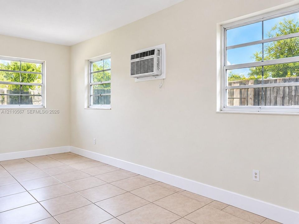 Neutral-toned interior space featuring light-colored ceramic tile flooring, three multi-pane windows, and a wall-mounted air conditioning unit