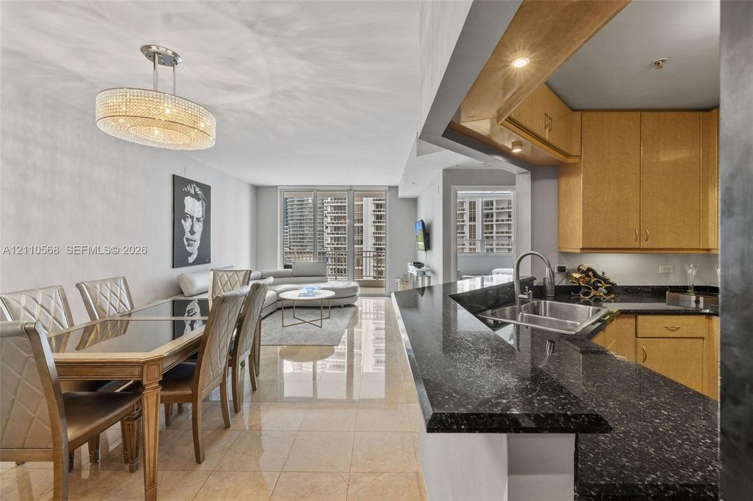 Open-concept living space featuring high-gloss tile flooring, kitchen with dark stone-finish countertops, custom light-toned wood cabinetry, stainless steel undermount sink, and a multi-tier crystal chandelier