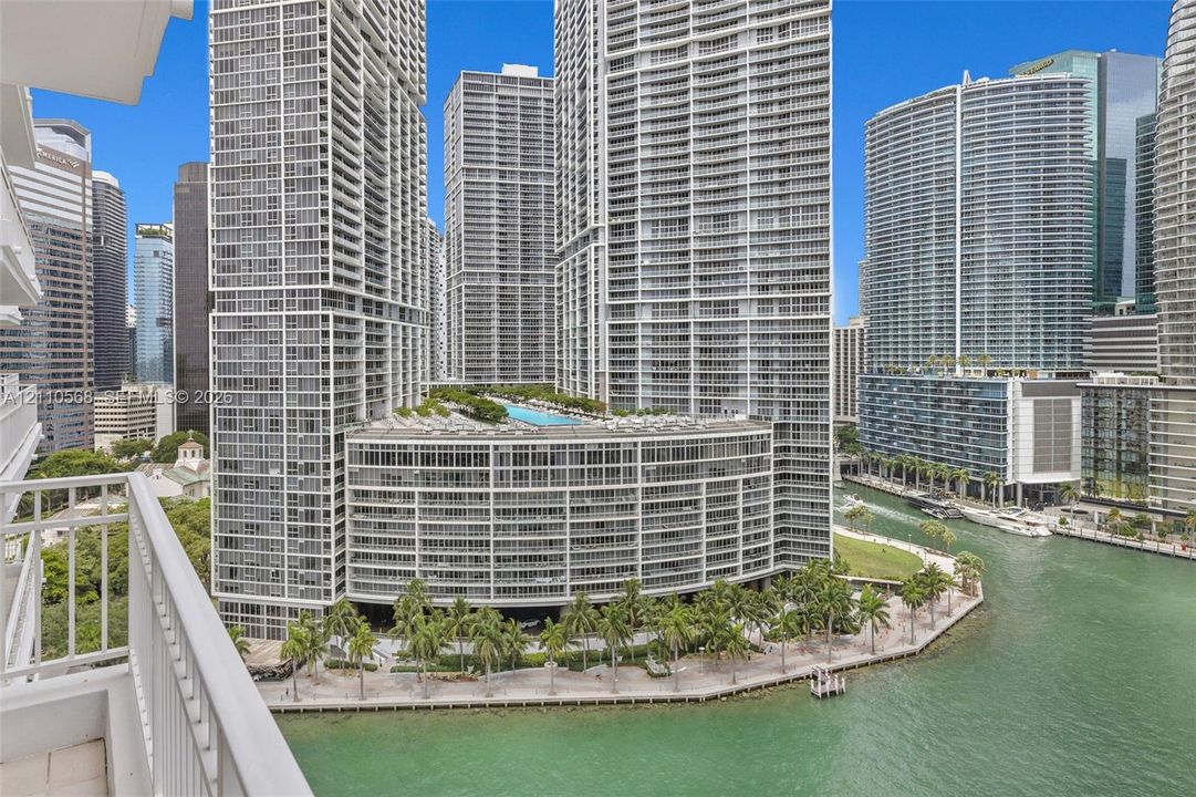 High-rise balcony with white metal railing overlooking a waterfront promenade, palm tree landscaping, and a multi-level complex featuring an elevated swimming pool