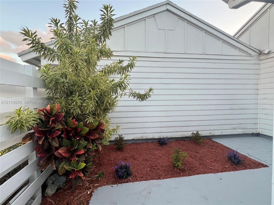Exterior space featuring white horizontal lap siding, decorative gable trim, a white privacy fence, painted concrete patio, and a garden bed with red mulch and ornamental shrubbery