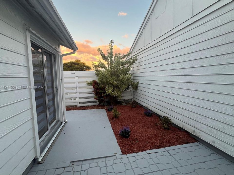 Exterior space featuring horizontal siding, a sliding glass door, a paved concrete patio, and a landscaped garden bed with mulch and plantings