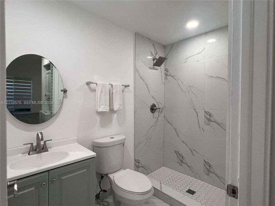 Bathroom featuring a vanity with a white countertop and oval under-mount sink, circular wall-mounted mirror, walk-in shower with large-format veined tile surround, hexagonal tile floor, and brushed-nickel hardware
