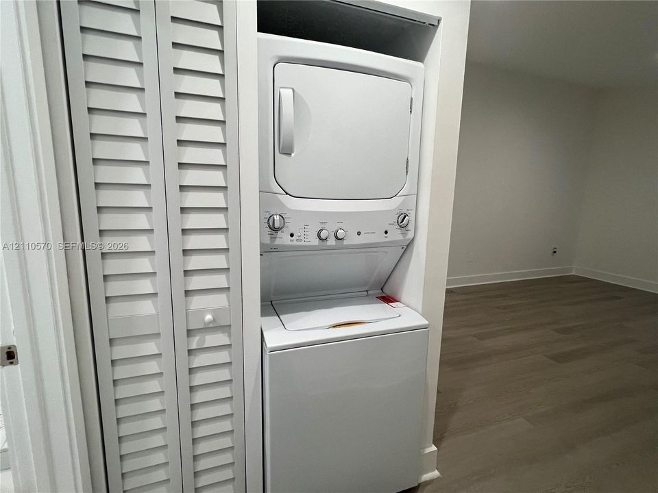 Stacked laundry appliance center behind white louvered bifold doors, featuring neutral-toned wood-finish flooring
