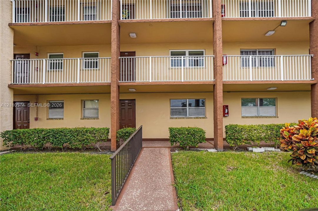 Multi-level exterior featuring stucco siding, metal balcony railings, a paved pedestrian walkway, and manicured shrubbery