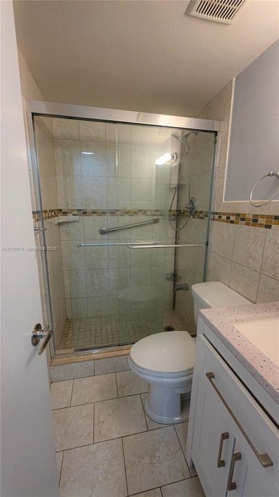 Walk-in shower featuring a sliding glass enclosure, mosaic tile accent border, and adjustable handheld showerhead, accompanied by a white vanity with a speckled countertop
