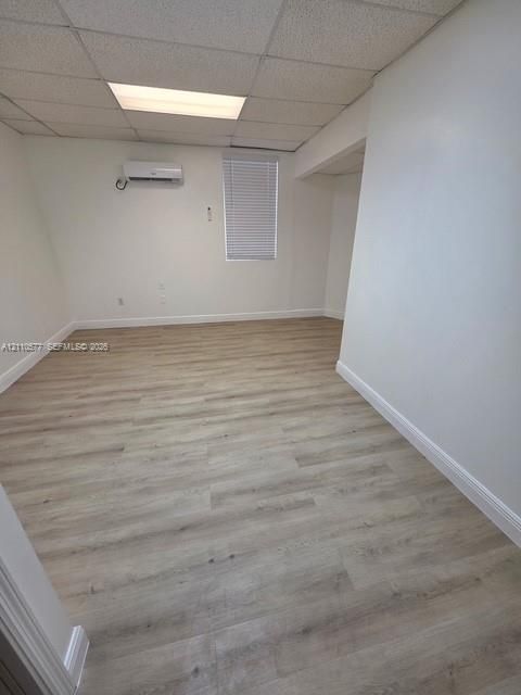 Interior room featuring wood-finish flooring, a wall-mounted ductless mini-split system, recessed grid ceiling with integrated lighting, and a single window with blinds