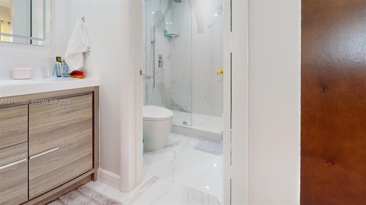 Modern vanity with wood-finish cabinetry, white stone-surround shower with glass enclosure, wall-mounted faucet hardware, and large-format tile flooring