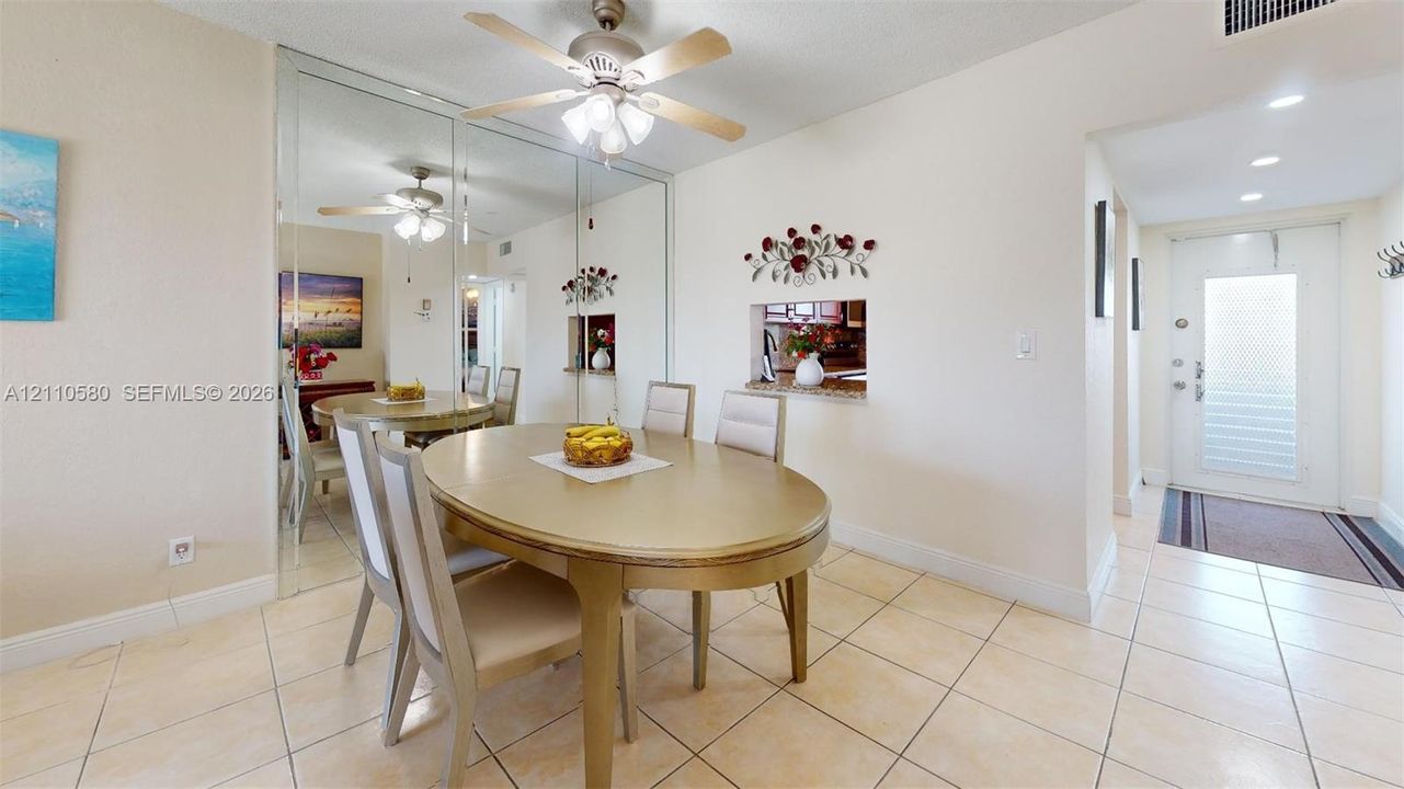 Dining area featuring large-format ceramic floor tiling, floor-to-ceiling mirrored wall paneling, a multi-light ceiling fan, and a pass-through opening to the kitchen