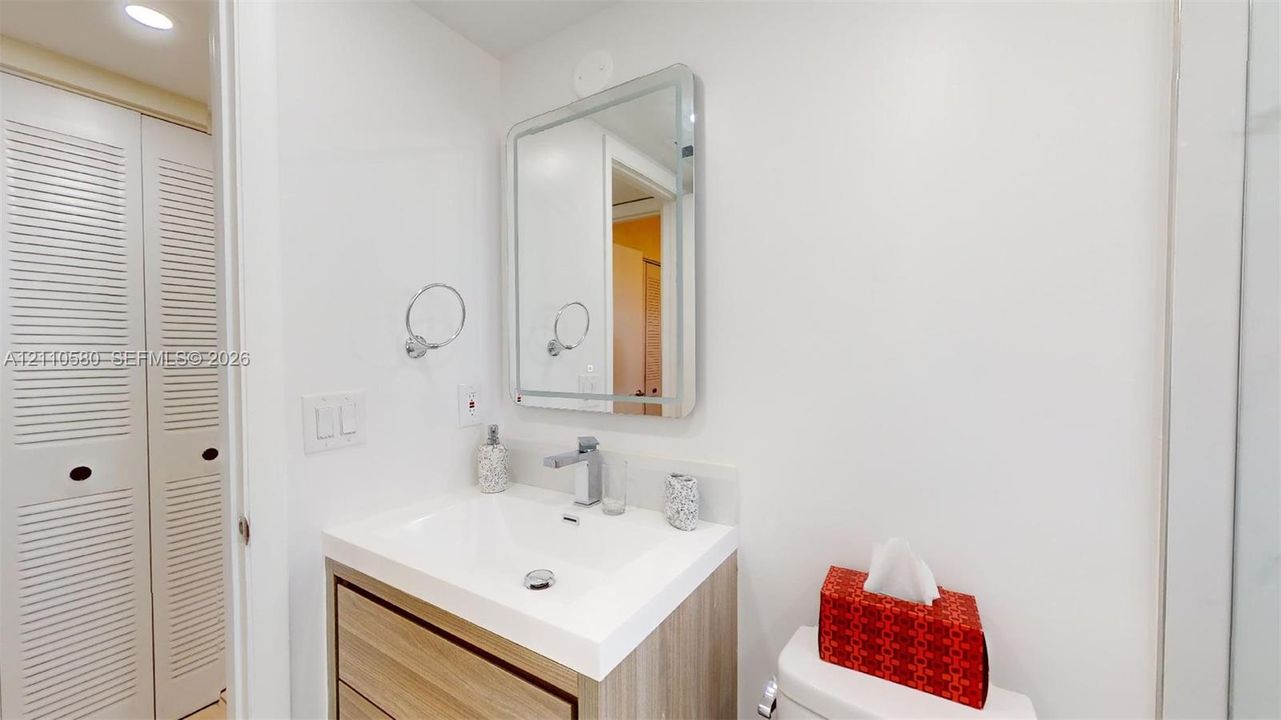 Bathroom featuring a wood-finish vanity with a white integrated basin, polished chrome faucet, rectangular wall-mounted mirror, and louvered bifold closet doors