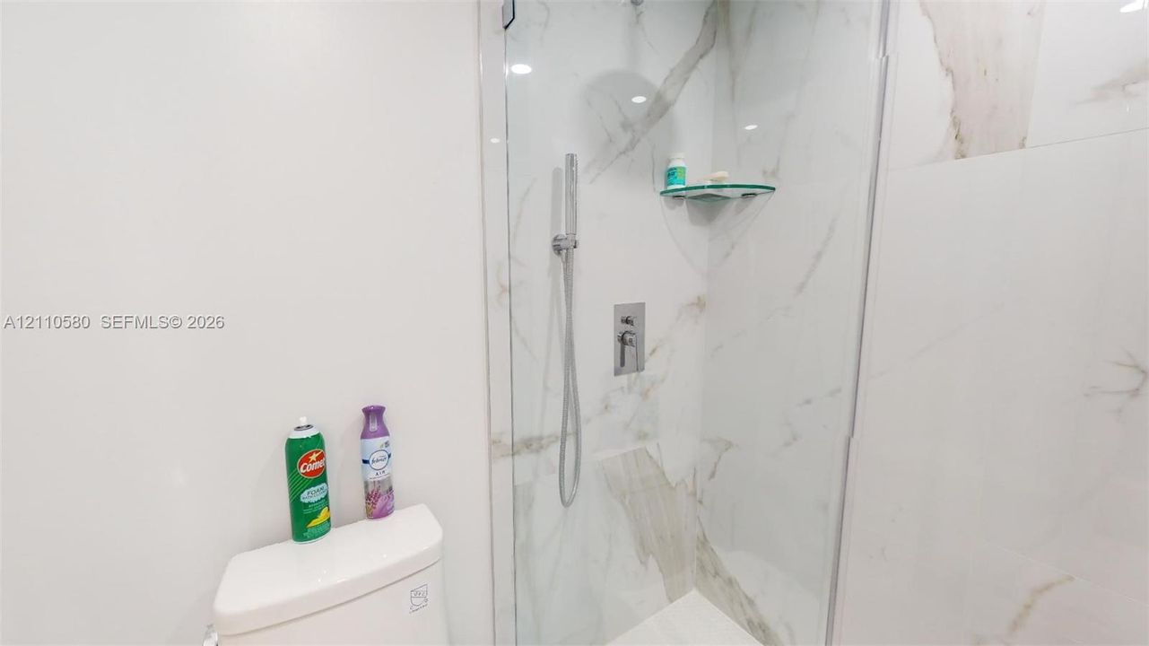 Walk-in shower featuring floor-to-ceiling large-format tile surround, glass enclosure, built-in corner shelving, and a wall-mounted handheld shower fixture