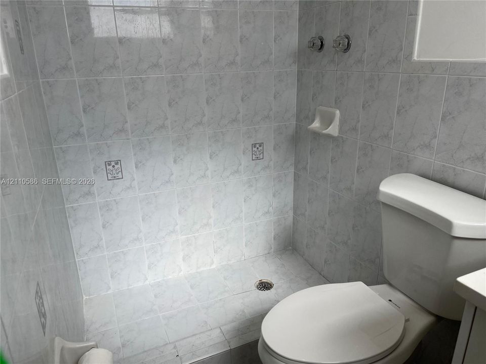Bathroom featuring floor-to-ceiling gray-toned tile, a built-in recessed soap dish, wall-mounted shower controls, and a standard-height toilet