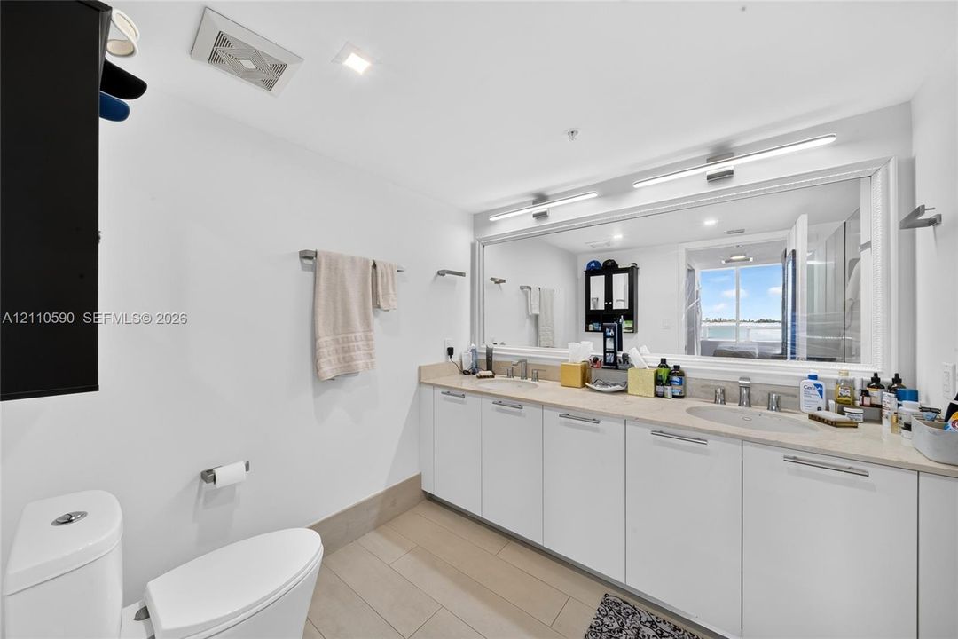 Double vanity with clean-lined white cabinetry, integrated sinks, stone-surface countertop, and a full-width framed wall mirror with integrated lighting