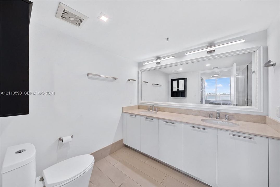 Dual-sink vanity with white cabinetry, light-toned stone-finish countertops, full-width framed mirror, integrated lighting, and tile flooring