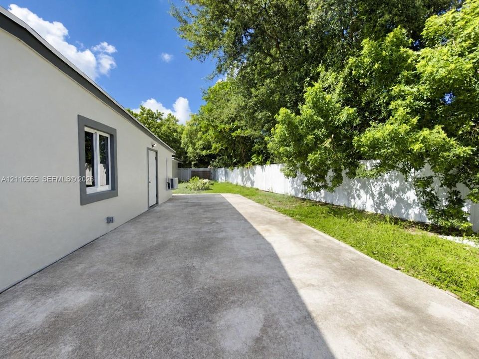 Concrete patio area featuring an exterior wall with smooth-finish siding, a window with grey trim, a solid exterior door, and perimeter fencing with mature trees