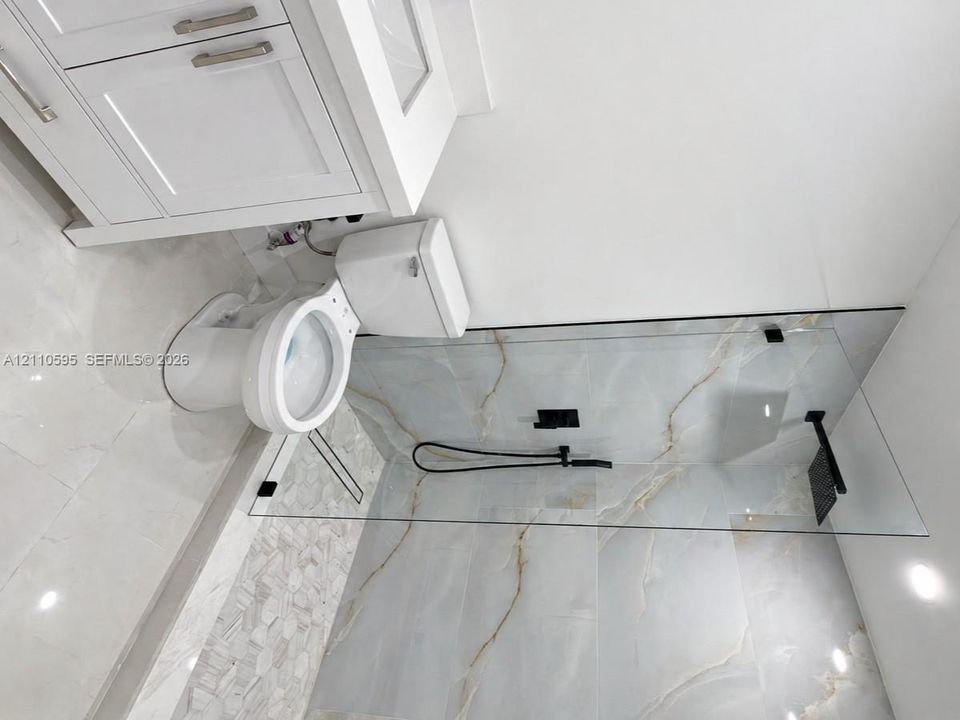 White cabinetry with brushed nickel hardware, light-toned large-format tile flooring, walk-in shower with frameless glass enclosure, matte black plumbing fixtures, and patterned decorative tile accents
