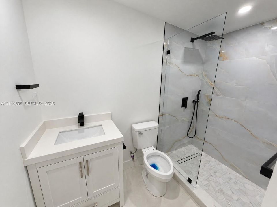 Bathroom featuring a vanity with a solid-surface countertop, walk-in shower with large-format tile walls, frameless glass enclosure, matte black rainfall shower head, and linear drain