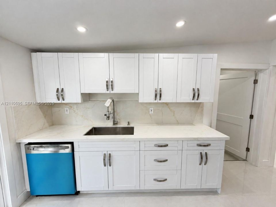 Kitchen featuring white Shaker-style cabinetry, light-toned stone-finish backsplash and countertops, undermount stainless steel sink with high-arc faucet, and integrated stainless steel dishwasher
