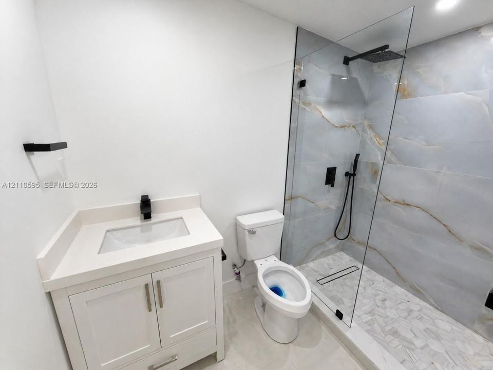 Bathroom featuring a white vanity with a composite countertop, matte black fixtures, a walk-in shower with large-format tile surround, and a frameless glass panel