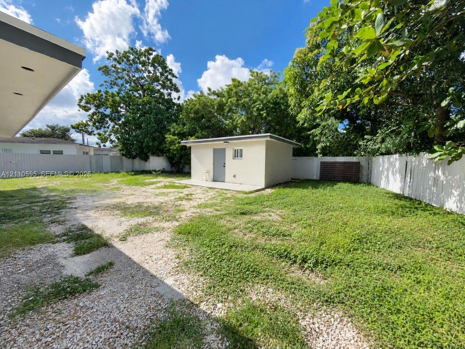 Expansive backyard featuring a detached storage structure, concrete pad, white vinyl perimeter fencing, and mature trees