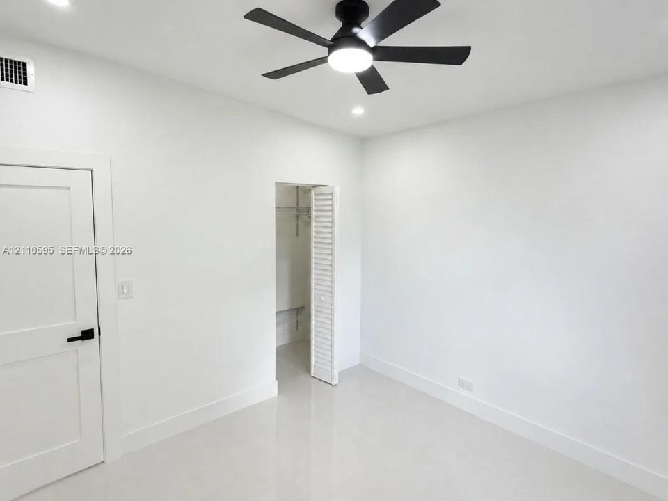 Neutral-toned interior featuring smooth-finish flooring, a matte black ceiling fan with integrated lighting, recessed ceiling lights, a solid-core panel door with matte black hardware, and a closet with louvered doors