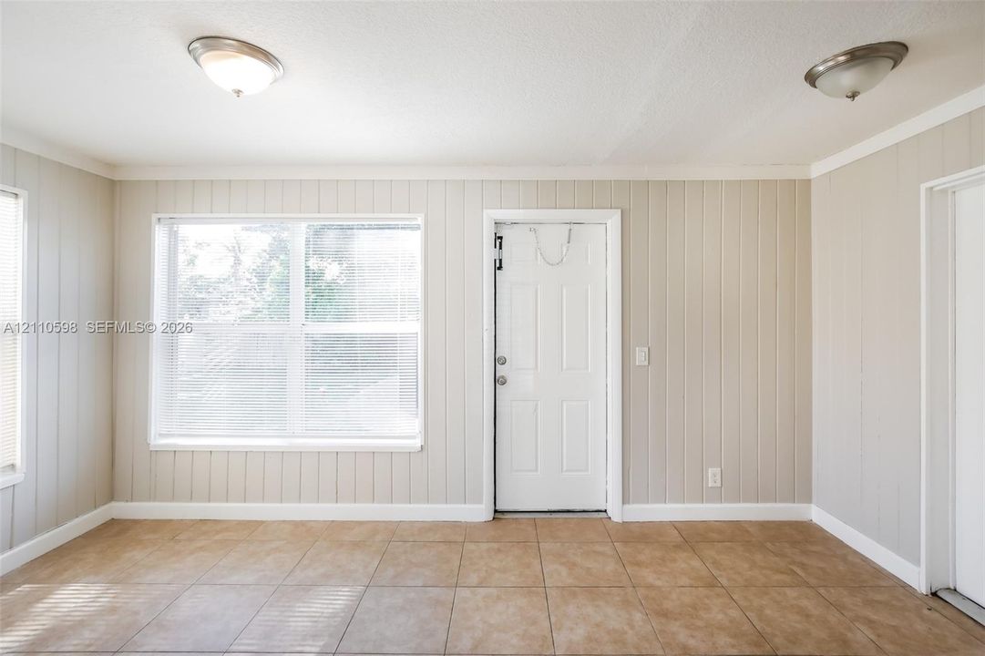 Interior space featuring neutral-toned vertical paneled walls, large format ceramic tile flooring, two flush-mount ceiling light fixtures, and a white multi-pane exterior entry door