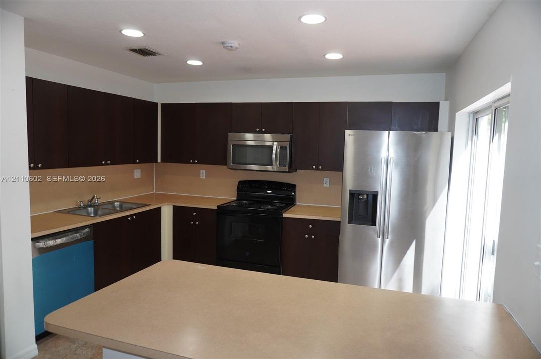 Kitchen featuring dark wood cabinetry, solid surface countertops, stainless steel refrigerator, over-the-range microwave, and recessed lighting