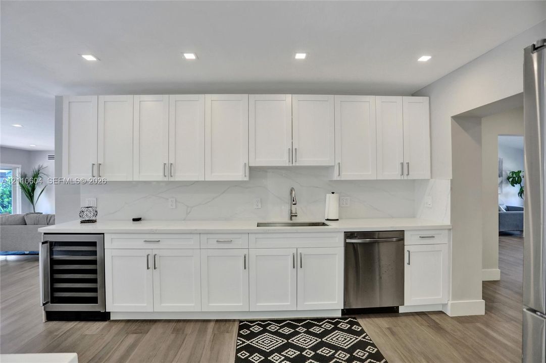 White shaker-style cabinetry, solid-surface countertop with integrated backsplash, stainless steel under-mount sink with chrome faucet, built-in beverage refrigerator, and wood-finish flooring