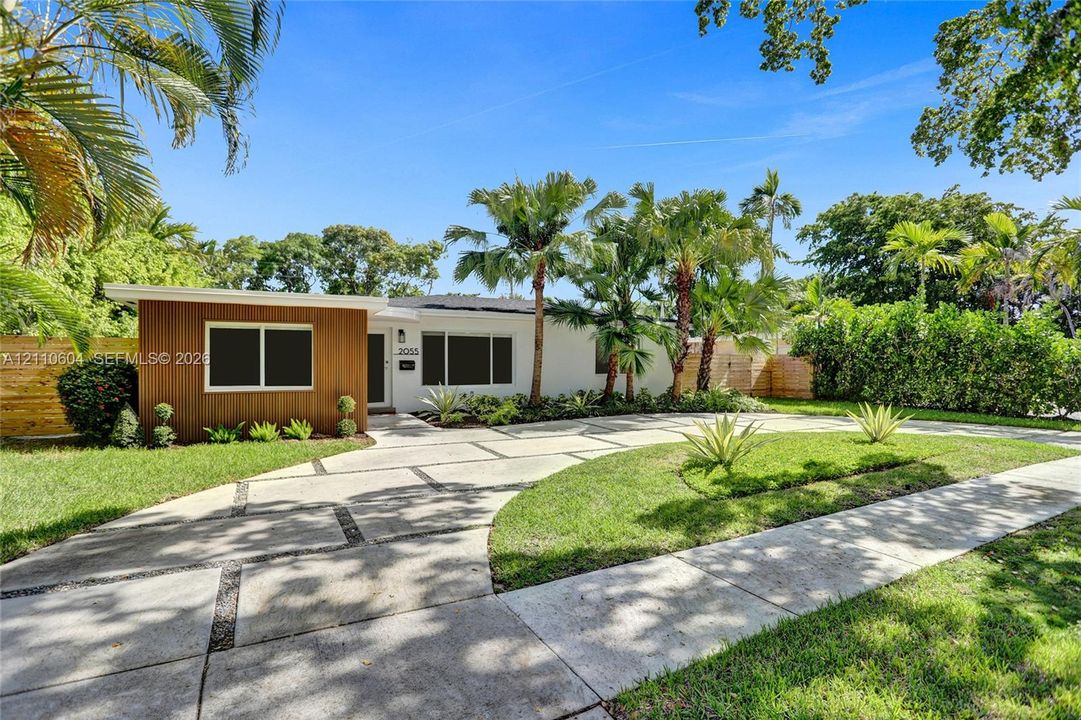 Mid-century modern exterior featuring wood-panel accent siding, smooth white stucco finish, concrete paver driveway with aggregate inlay, and mature palm landscaping