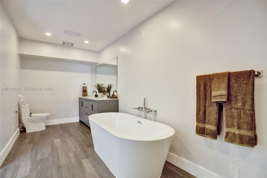 Freestanding white soaking tub with wall-mounted chrome filler, gray vanity cabinet, rectangular mirror, recessed lighting, and wood-finish flooring