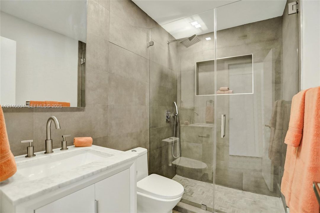 White vanity with stone-finish countertop, undermount sink, and brushed-nickel faucet, accompanied by a walk-in shower featuring large-format tile walls, glass enclosure, built-in shelving niche, and rain-style showerhead