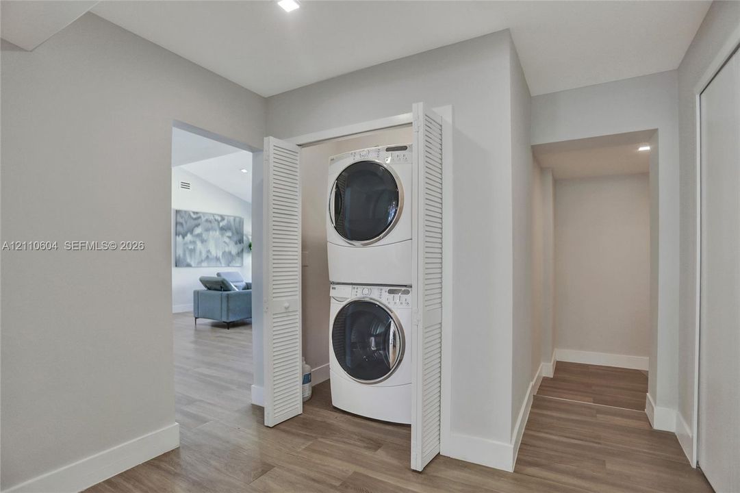 Laundry alcove featuring a stacked washer and dryer unit enclosed by white louvered bifold doors, complemented by wood-finish flooring and recessed ceiling lighting