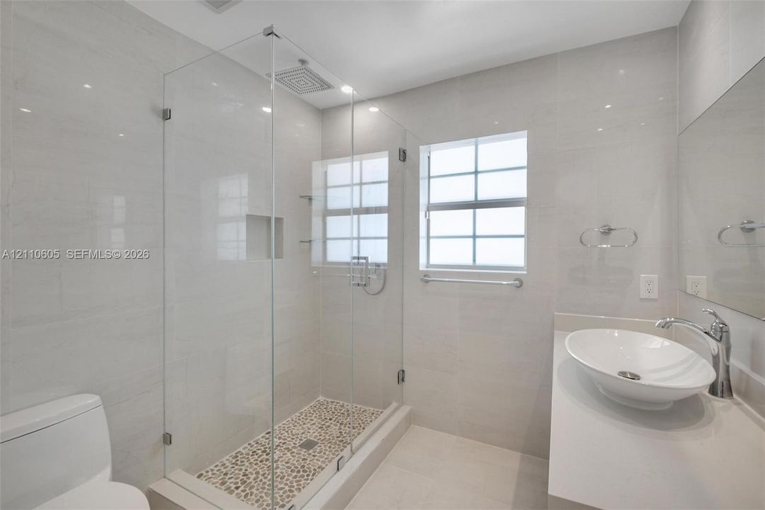 Bathroom featuring a glass-enclosed walk-in shower with pebble-stone flooring, a wall-mounted rainfall showerhead, a quartz-topped vanity with an oval vessel sink, and large-format porcelain wall tile
