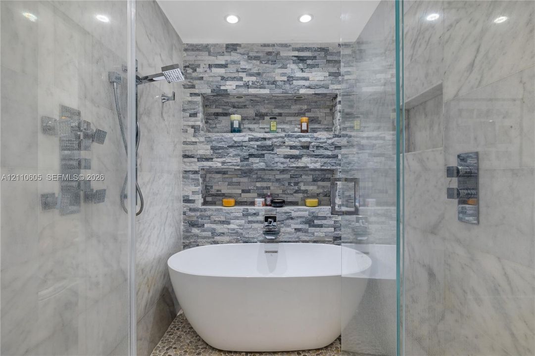Freestanding soaking tub with polished chrome fixtures, integrated stacked-stone feature wall with dual recessed shelving, stone pebble flooring, and frameless glass shower enclosure