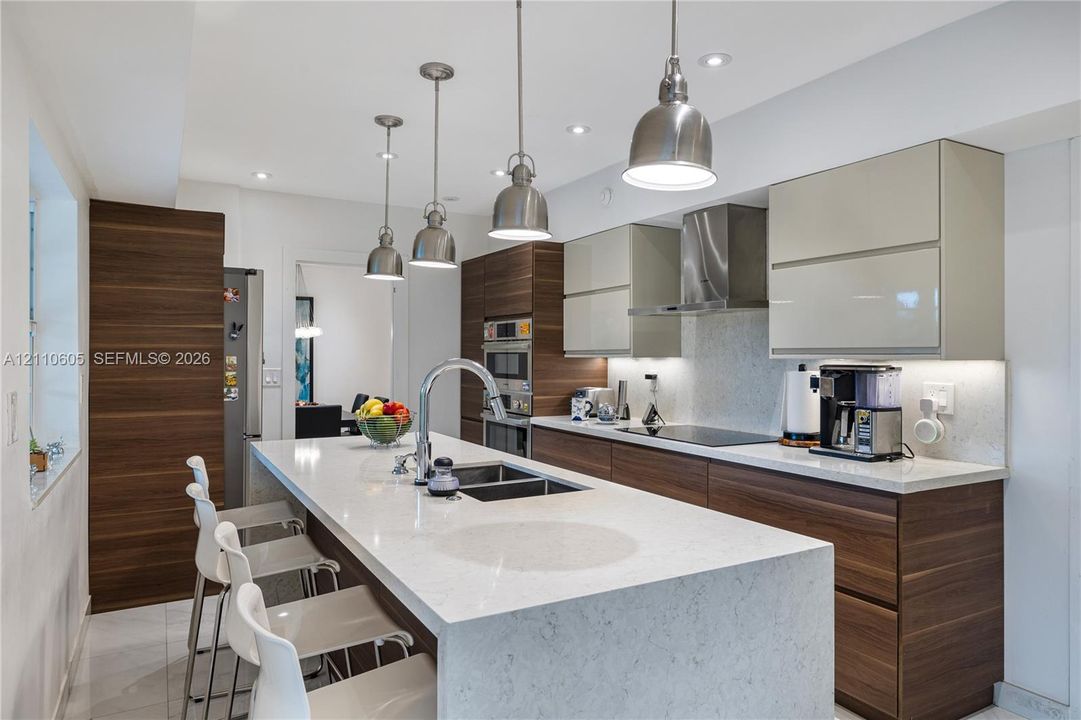 Kitchen featuring a waterfall-edge center island, wood-finish cabinetry, light-toned solid surface countertops, stainless steel pendant lighting, and a stainless steel wall-mounted range hood