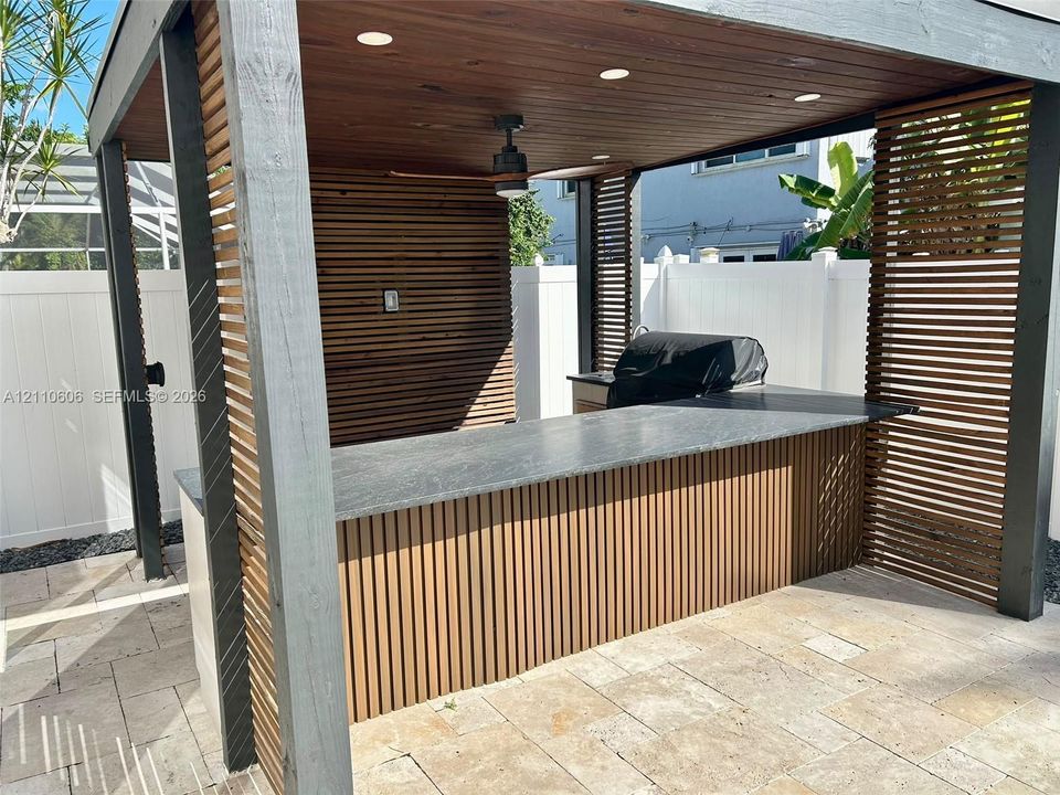 Covered outdoor kitchen featuring horizontal wood slat privacy walls, stone-finish bar top, recessed lighting, and a ceiling-mounted fan, situated on a stone paver patio