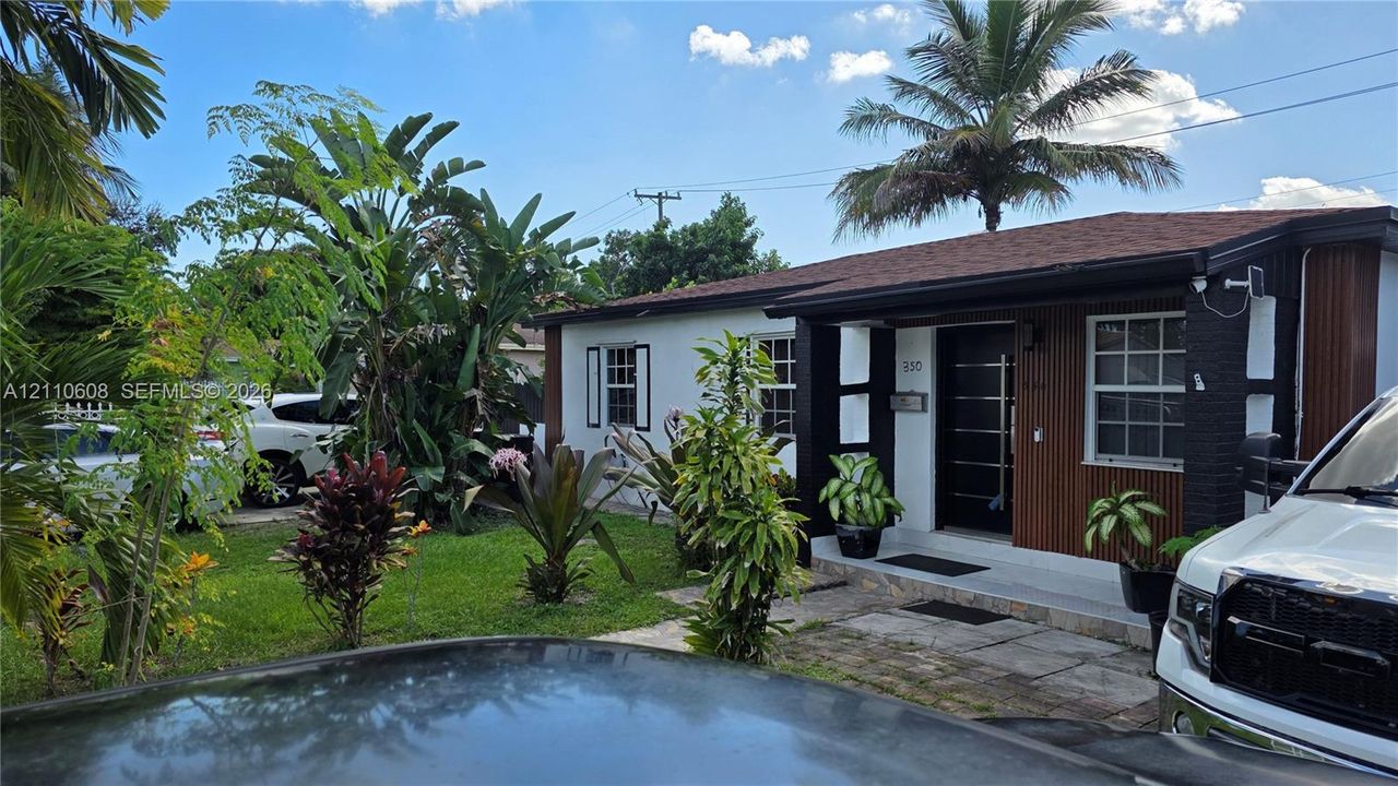 Exterior facade featuring mixed-material siding, a covered entryway, multi-panel front door, and established tropical landscaping