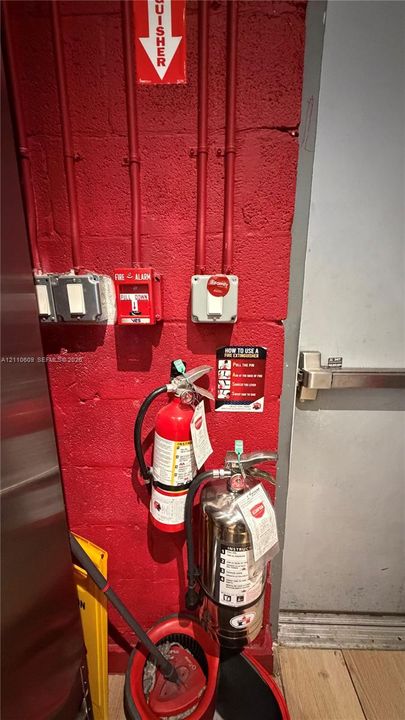 Painted red masonry wall featuring fire suppression equipment, including a fire alarm pull station, two mounted fire extinguishers, and a commercial-grade metal exit door with a crash bar