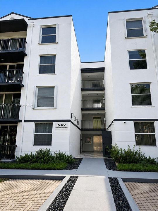 Modern multi-story facade with clean-lined exterior, vertical architectural interest, dark metal balconies, and geometric paved hardscaping