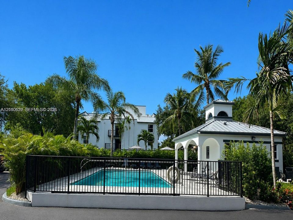 Swimming pool with metal perimeter fencing, covered cabana featuring arched architectural detailing and a metal roof, surrounded by mature palm trees and manicured landscaping