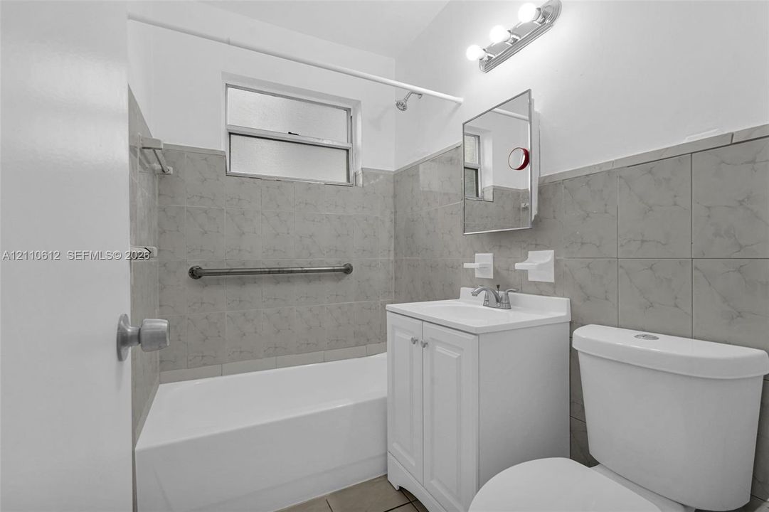 Bathroom featuring a white vanity cabinet, rectangular sink with a chrome faucet, light-toned ceramic wall tiling, shower-tub combination, and three-bulb vanity lighting