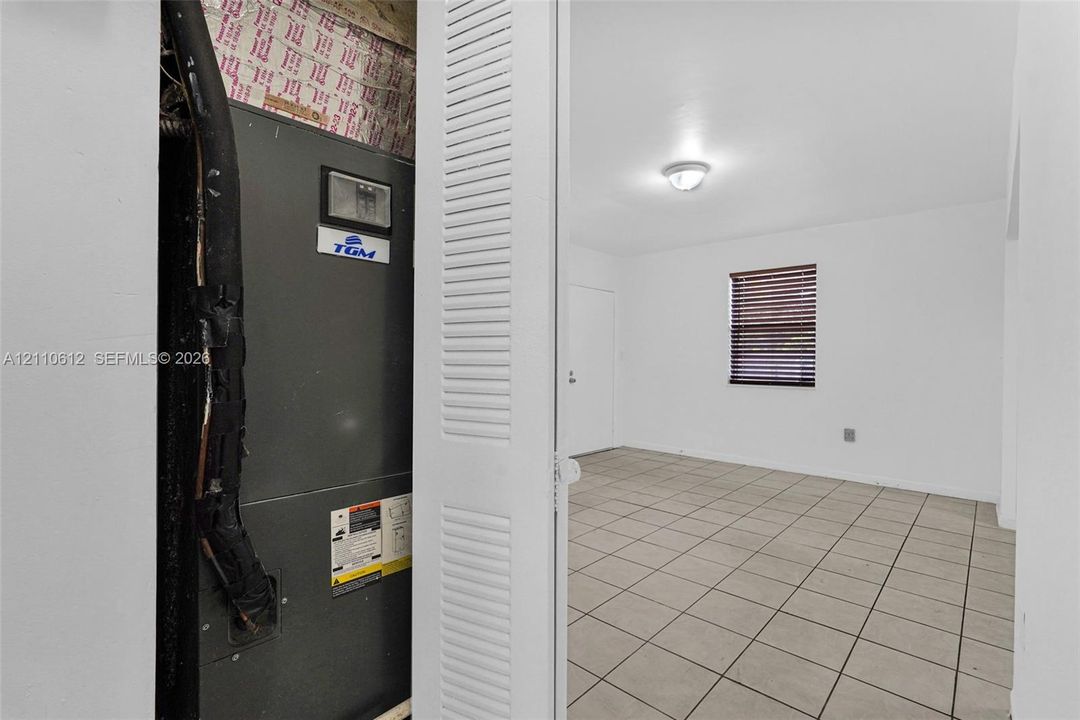 Utility closet containing a TGM HVAC unit, paired with a room featuring neutral tile flooring, a window with horizontal blinds, and a flush-mount ceiling light fixture